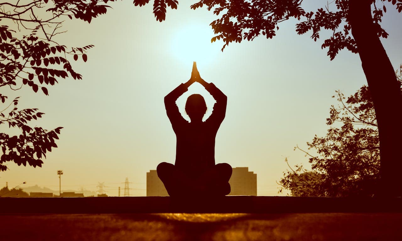 Silhouette of a person meditating outdoors at sunrise, hands joined above the head
